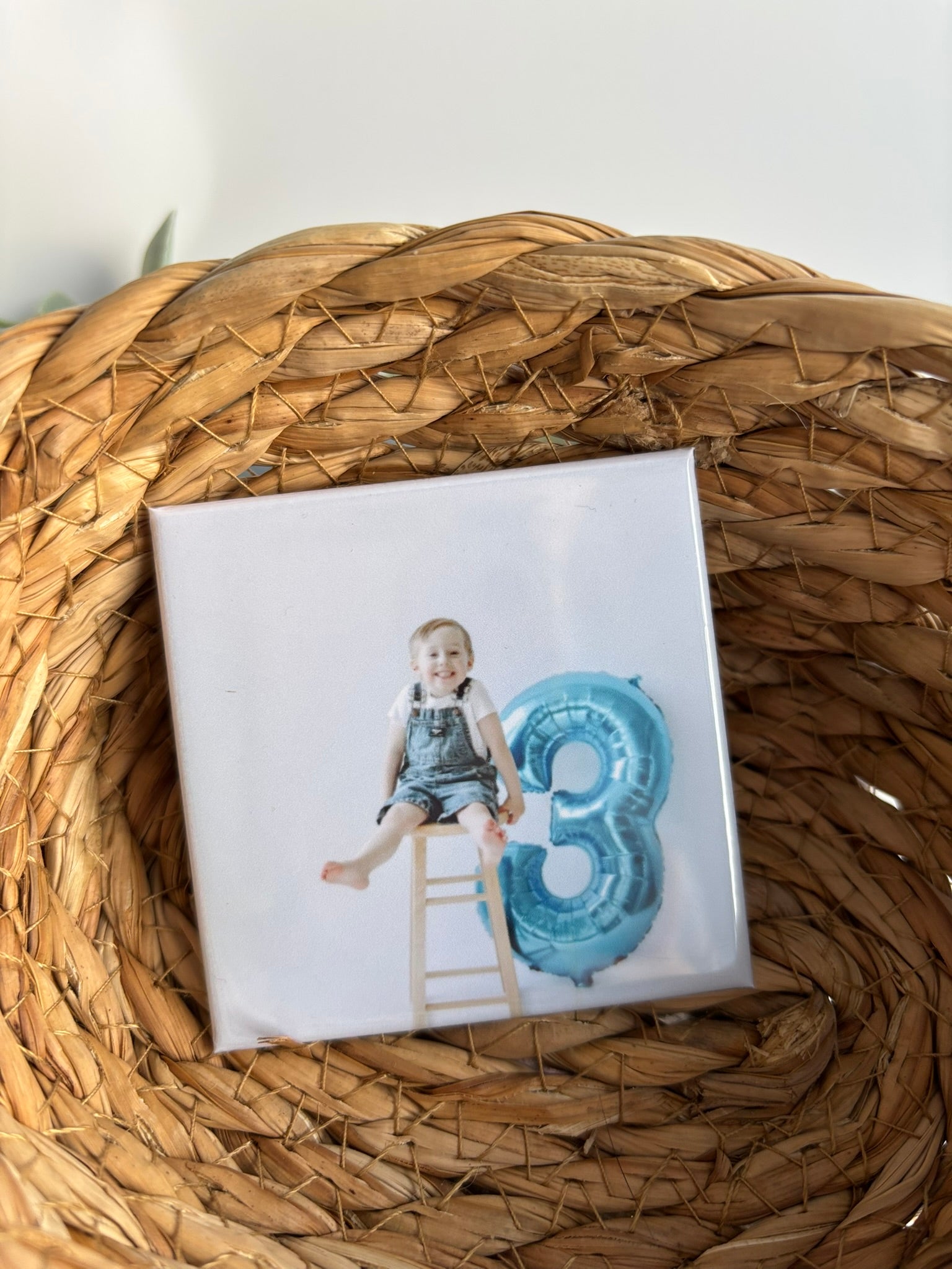 Square magnet with a child on a chair and a blue balloon number '3', celebrating third birthday on a woven surface