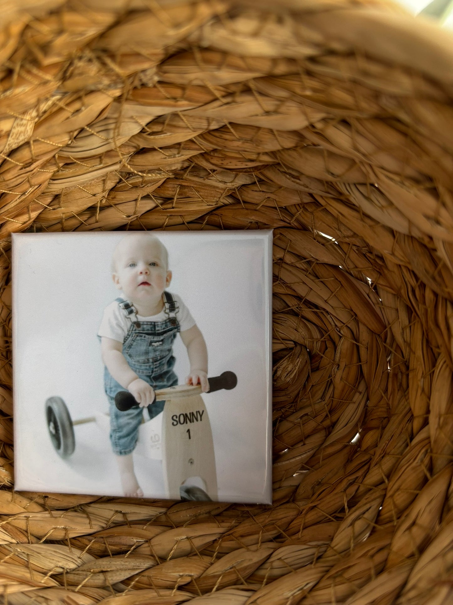 Square photo print of a child on a tricycle first birthday photoshoot placed on woven straw.